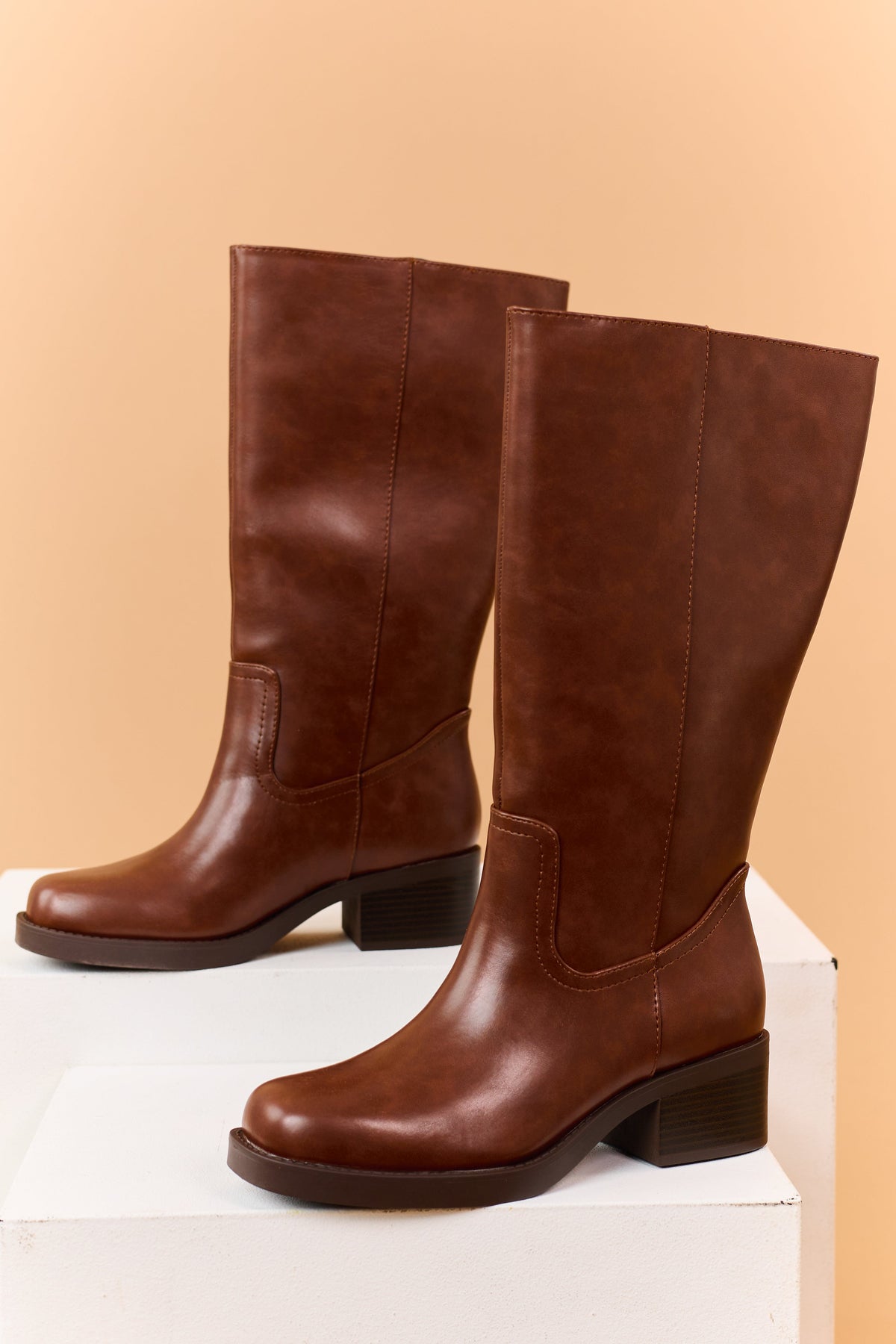 Pair of brown knee-high boots displayed on a white pedestal.